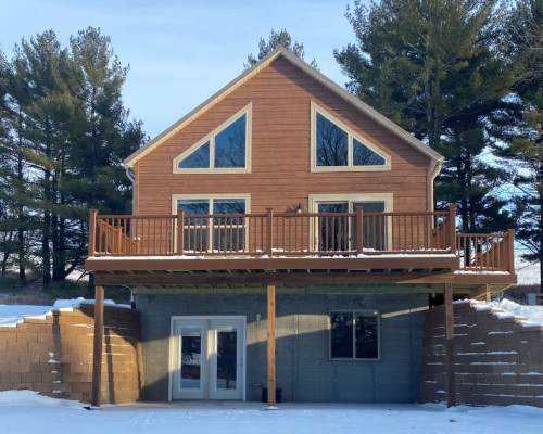 A two-story wooden house with large windows, a deck, and snow-covered ground, surrounded by trees.