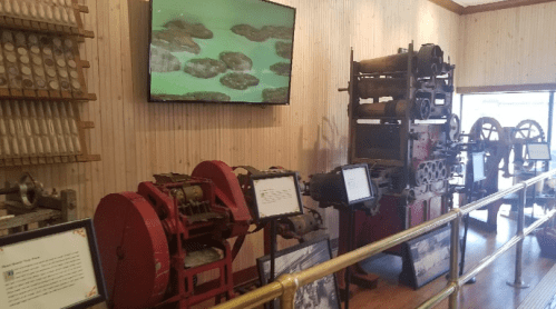 Historic machinery displayed in a museum, featuring various printing presses and a screen showing animated content.