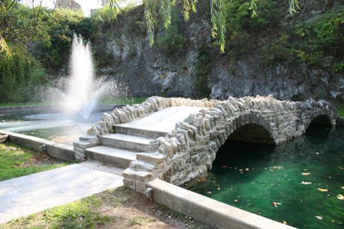 A stone bridge arches over a tranquil pond with a fountain, surrounded by lush greenery and rocky cliffs.
