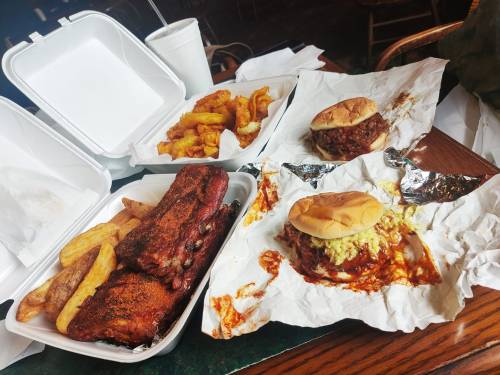 A table with takeout containers featuring ribs, fries, and a pulled pork sandwich, all wrapped in paper.