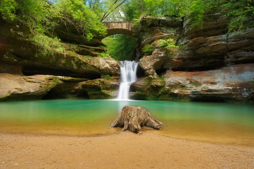 A serene waterfall cascades into a turquoise pool, surrounded by lush greenery and a stone bridge in the background.