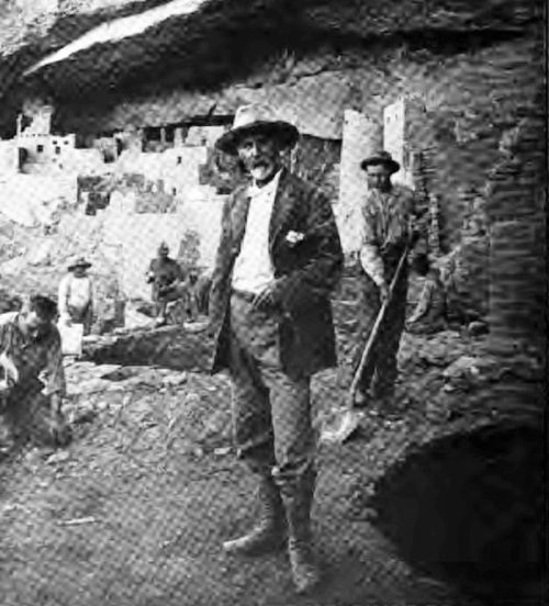 A group of men, including one in a suit and hat, stands near a cliffside dwelling, with tools and excavation sites visible.