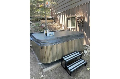A wooden hot tub with a cover, accompanied by a small set of black and white steps, set against a house exterior.