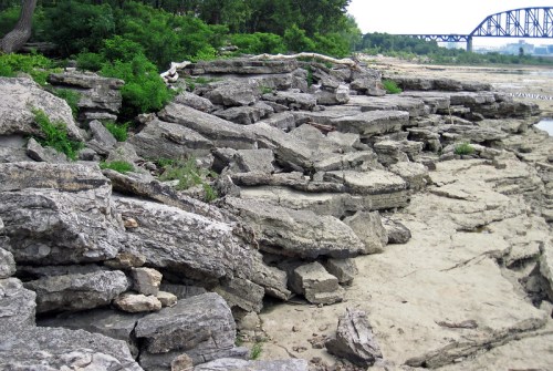 Rocky shoreline with large stone formations and sparse greenery, under a bridge in the background.