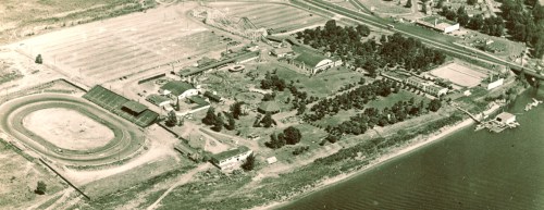 Aerial view of a park with a racetrack, amusement rides, and green spaces near a river.