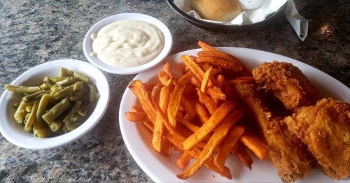 A plate of fried chicken, sweet potato fries, green beans, and a side of creamy dipping sauce on a textured surface.