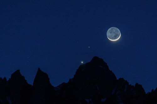A crescent moon shines brightly over rugged mountain peaks against a deep blue night sky, with stars twinkling above.