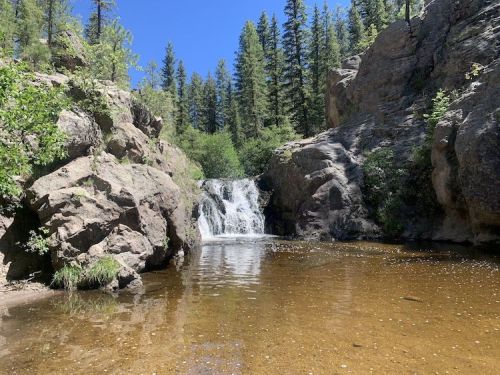 A serene waterfall cascades into a clear pool, surrounded by rocky cliffs and lush green trees under a bright blue sky.