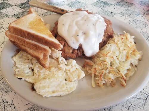 A plate of fried chicken topped with white gravy, served with toast, scrambled eggs, and hash browns.