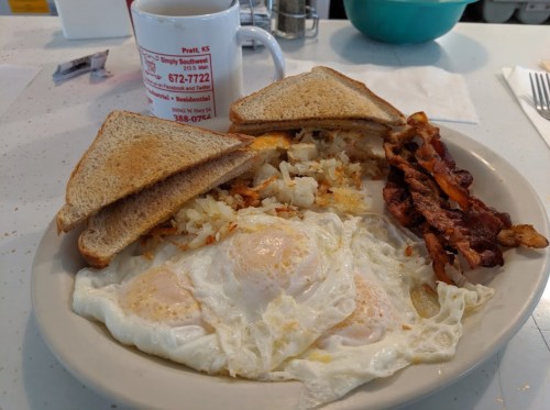 A plate with two fried eggs, bacon, hash browns, and toast, alongside a coffee cup on a diner table.