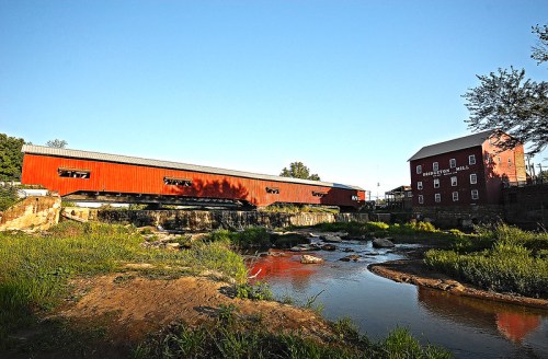 A red covered bridge spans a stream, with a historic mill building nearby, surrounded by greenery and clear blue skies.