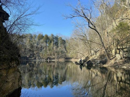 A serene river scene with trees and rocky cliffs, reflecting in calm water under a clear blue sky.