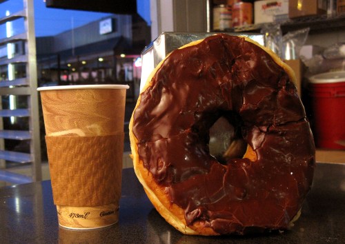 A large chocolate frosted donut next to a coffee cup on a countertop, with a blurred background.