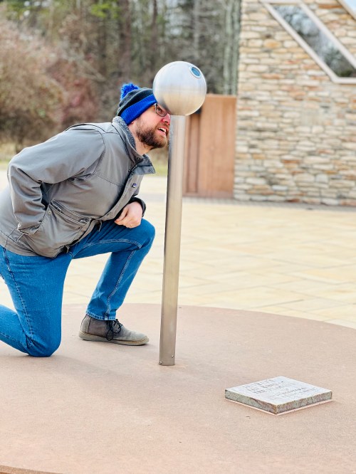 A person kneels beside a metal pole with a spherical top, looking through it in an outdoor setting.