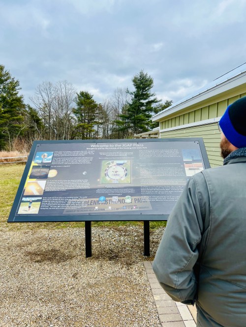 A person stands in front of an informational sign about a park, surrounded by trees and a cloudy sky.