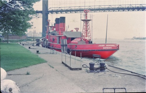 A red boat named "John Kendall" docked by a river, with a bridge in the background and green grass nearby.
