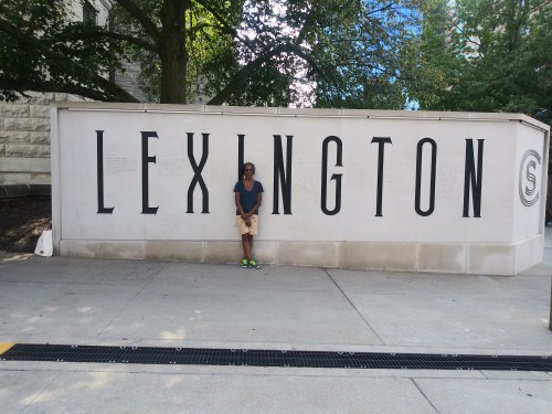 A person stands next to a large "LEXINGTON" sign on a wall, surrounded by trees and urban scenery.