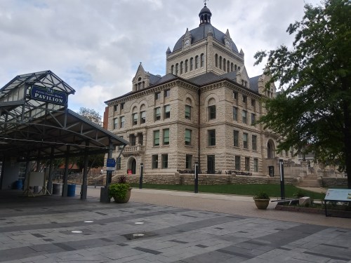 Historic building with a clock tower, surrounded by greenery, next to a modern pavilion in a public square.