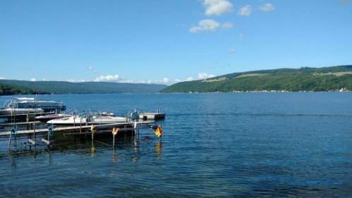 A serene lake view with boats docked, surrounded by green hills and a clear blue sky.