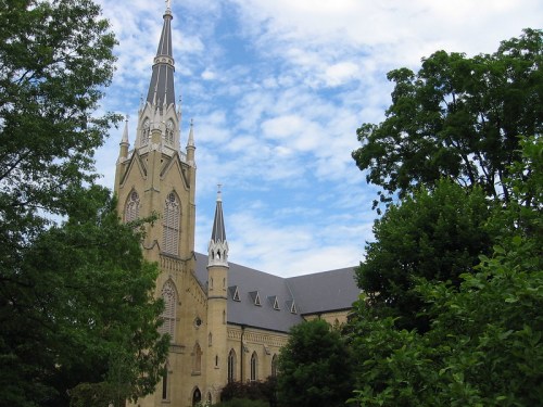 A tall church with a spire, surrounded by green trees and a blue sky with scattered clouds.