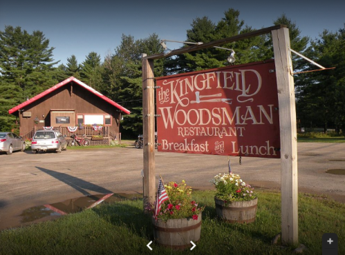 Sign for The Kingfield Woodsman Restaurant, featuring a rustic building and flower planters, surrounded by trees.