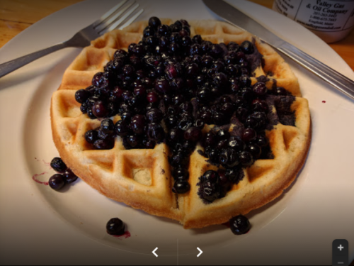 A golden waffle topped with a generous serving of fresh blueberries on a white plate. Fork and knife beside it.