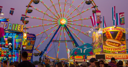A vibrant carnival scene with a Ferris wheel, food stalls, and colorful lights at dusk.