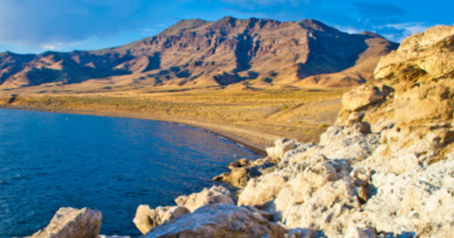 A rocky shoreline meets a calm blue lake, with mountains rising in the background under a clear sky.