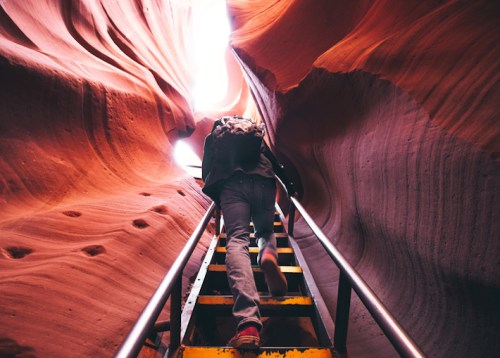 A person climbs stairs through a narrow, colorful canyon with smooth, curved rock walls. Light shines from above.