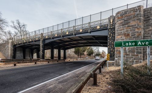 A decorative bridge spans over a road, with a sign for Lake Ave visible on the right. Trees line the background.