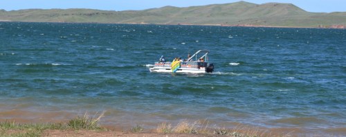 A small boat with passengers sails on a choppy blue lake, surrounded by green hills under a clear sky.