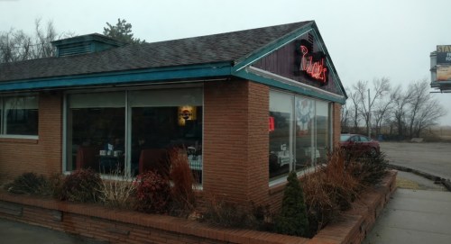 A brick diner with large windows, a neon sign, and a cloudy sky in the background.