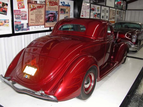 A shiny red vintage car displayed in a museum, with framed posters and photos on the wall behind it.