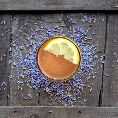 A glass of drink with a lemon slice, surrounded by dried lavender on a wooden surface.