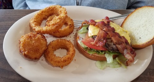 A plate with a cheeseburger topped with bacon, lettuce, and tomato, alongside crispy onion rings.