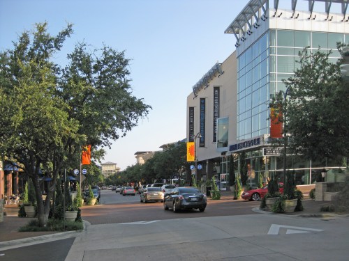 A street view featuring trees, modern buildings, and parked cars under a clear blue sky.