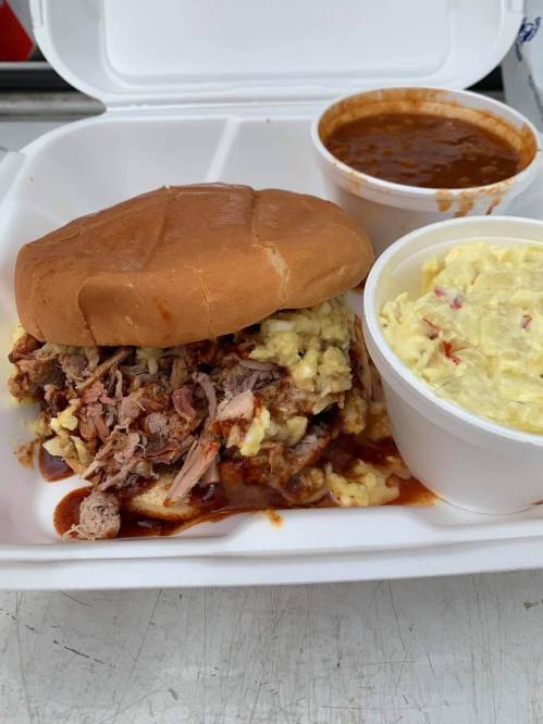 A pulled pork sandwich with mashed potatoes, served with a side of coleslaw and a cup of chili in a takeout container.