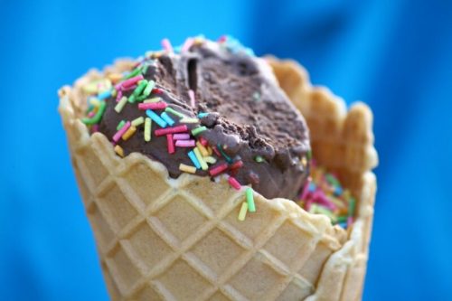 A close-up of a chocolate ice cream scoop in a waffle cone, topped with colorful sprinkles against a blue background.