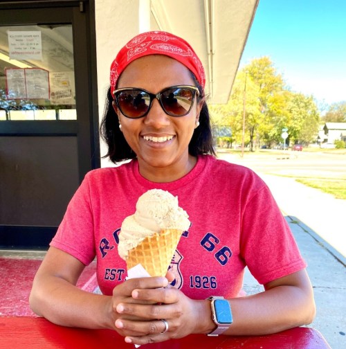 A smiling person wearing a red bandana and sunglasses holds a large ice cream cone while sitting at a table.