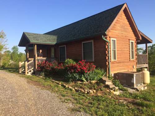 A wooden cabin with a green roof, surrounded by colorful flowers and a gravel path, under a clear blue sky.