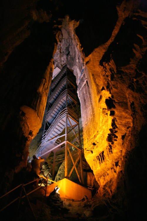 A narrow cave with a metal structure inside, illuminated by warm lighting, showcasing rugged rock formations.