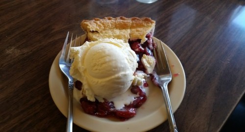 A slice of cherry pie topped with a scoop of vanilla ice cream on a plate, with two forks beside it.