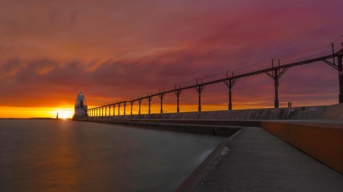 A serene sunset over a pier with a lighthouse, casting vibrant colors across the sky and reflecting on the water.