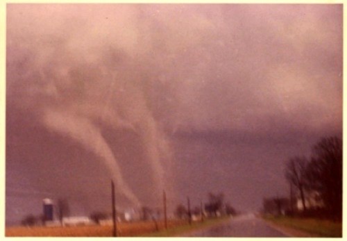 A dramatic sky with dark clouds and multiple tornadoes forming over a rural road and fields.