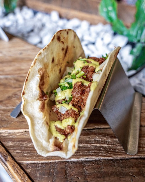 A taco filled with meat, avocado, and chopped vegetables, served in a metal holder on a wooden table.