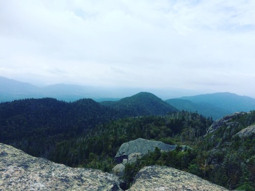 A panoramic view of lush green mountains under a cloudy sky, with rocky foreground and distant hills.