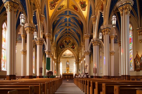 Interior of a grand church featuring ornate arches, colorful stained glass windows, and wooden pews.