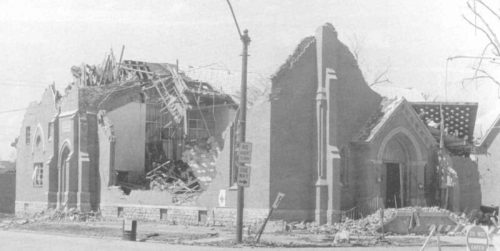 A damaged brick church with a collapsed roof and debris scattered outside, showing signs of significant destruction.