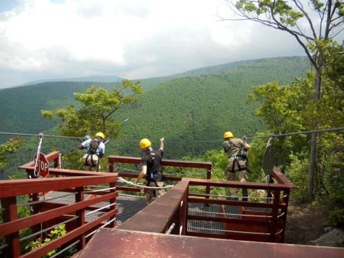Three people in helmets prepare to zip line over a lush green mountain landscape on a sunny day.