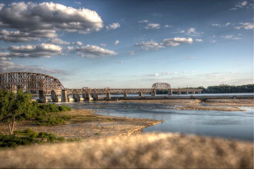 A river scene featuring a bridge, lush greenery, and a cloudy sky, with calm waters reflecting the landscape.